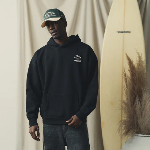 Person wearing the Huega House Trademark Hat in Natural and Green in a sunny outdoor or everyday casual setting